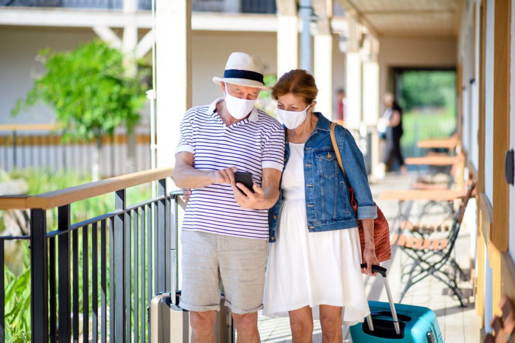 A couple, both wearing face masks, appear on the balcony of a hotel or accommodation with a suitcase. They are both looking at a phone.